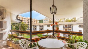Un balcón con una mesa y sillas blancas, unidades de aire acondicionado y vistas de edificios circundantes.