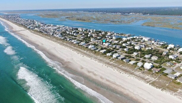 Vue aérienne d’une zone résidentielle côtière, avec des maisons le long de la plage et une jetée qui s’avance dans l’eau.