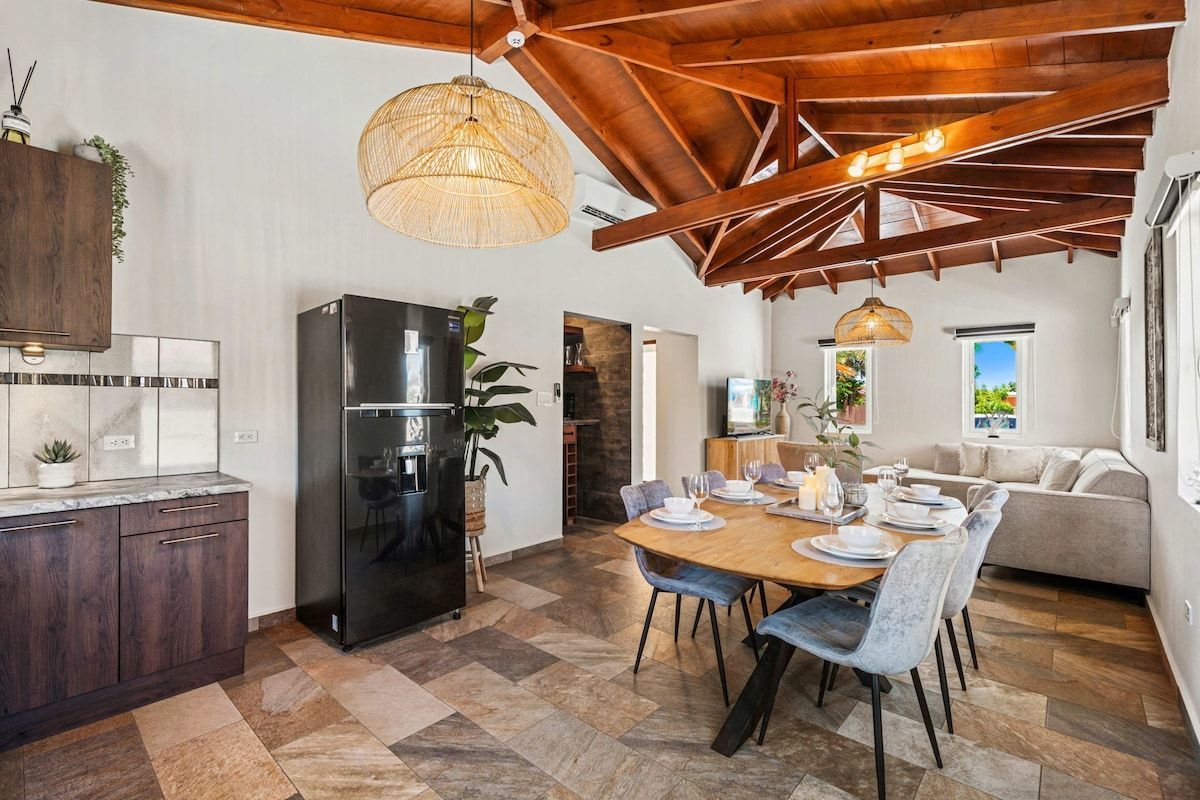 A modern kitchen with a built-in refrigerator, a dining area with a wooden table and chairs, and a high ceiling with exposed beams.