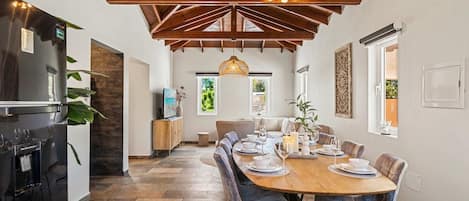 A dining area with a wooden table and chairs, a hanging light fixture, and a kitchen area with a refrigerator and cabinets.