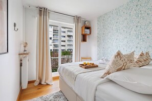 A bedroom with a bed, a tray with breakfast, a window with curtains, a wall shelf, and a view of a building. - Charming 2BR Flat Near Eiffel Tower & Invalides (Paris)