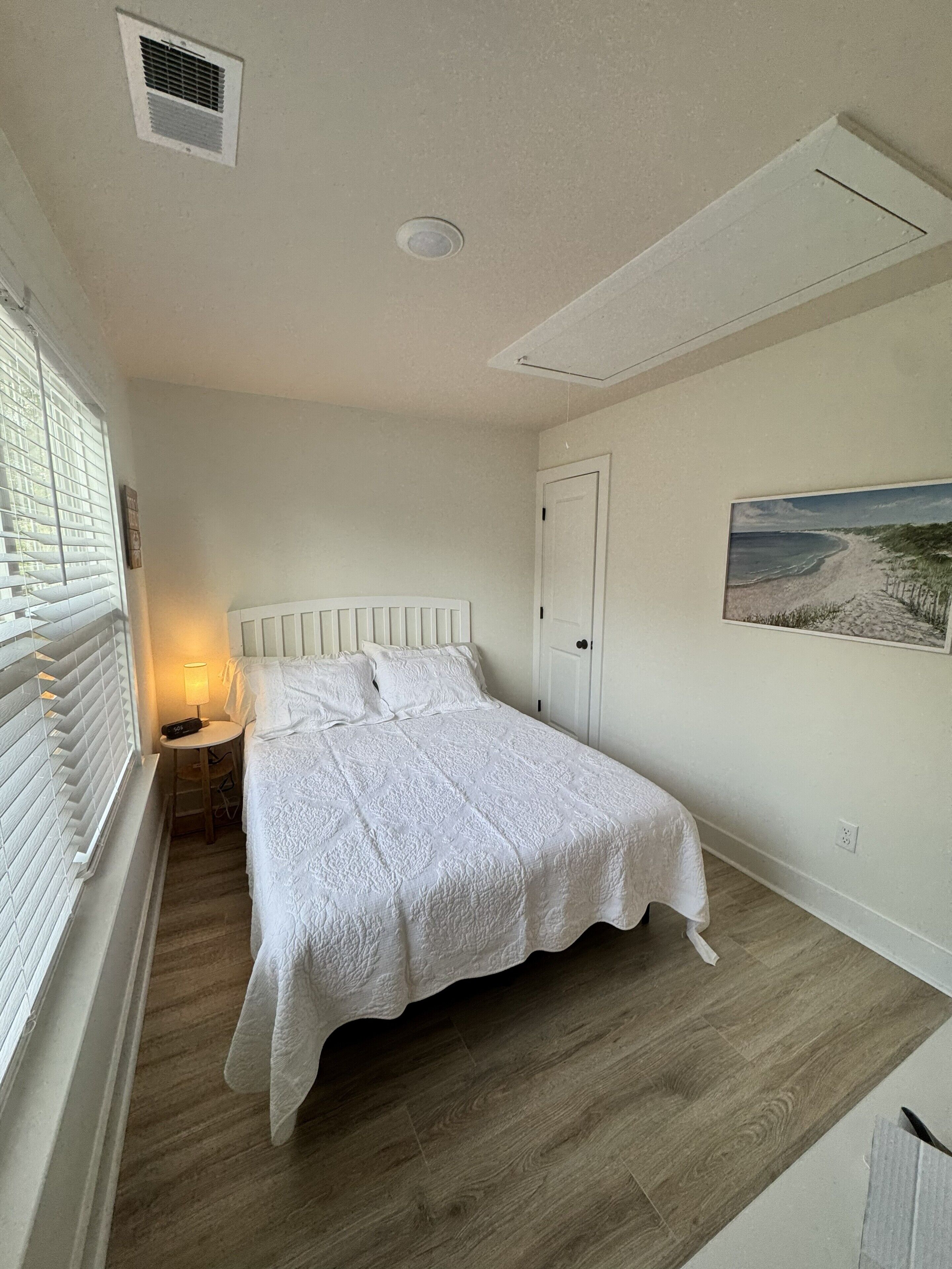 A bedroom with a bed, a nightstand, a lamp, a door, and a painting on the wall.