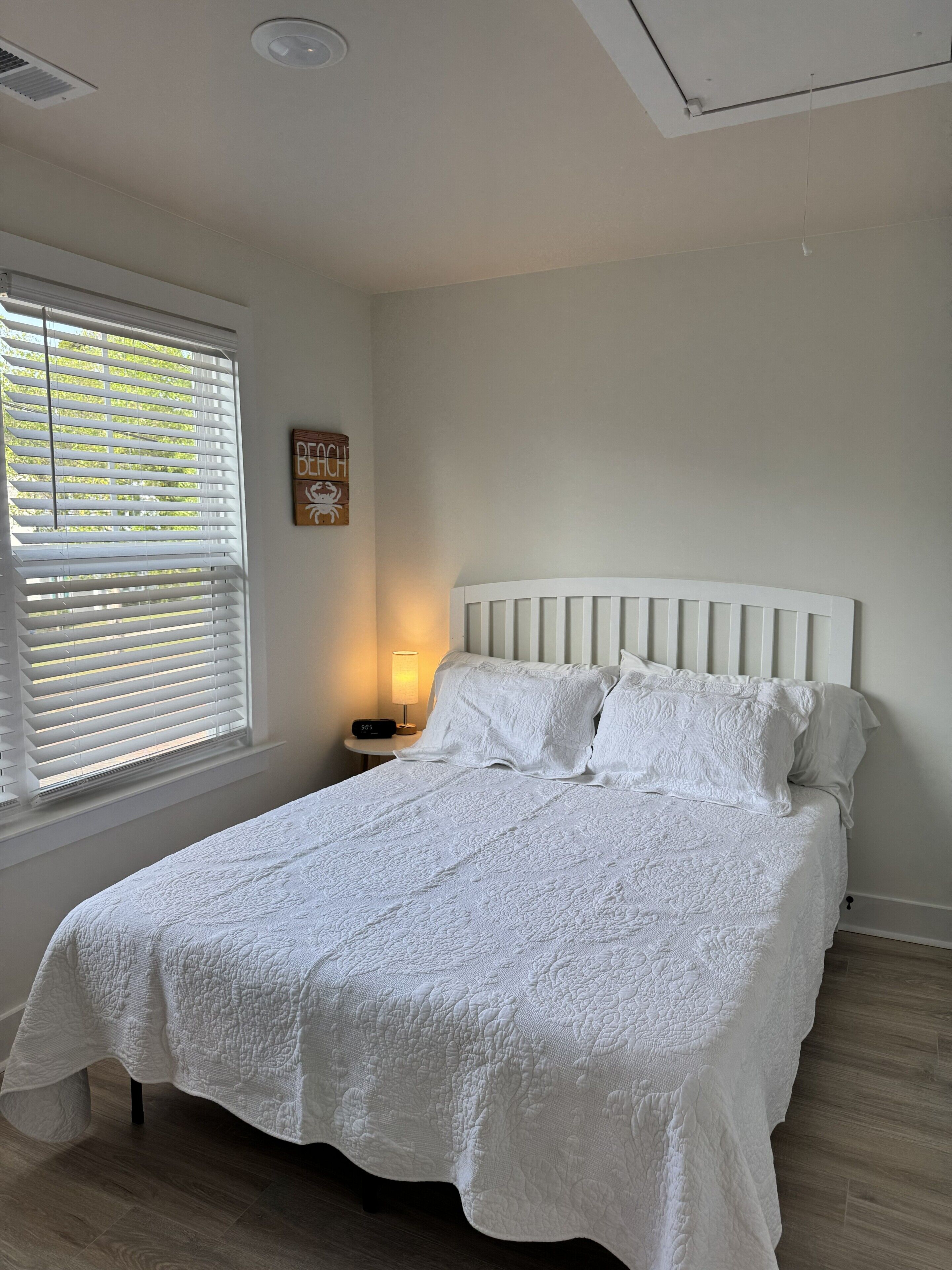 Un dormitorio con cama, mesita de noche, lámpara, ventana con persianas y un adorno en la pared.