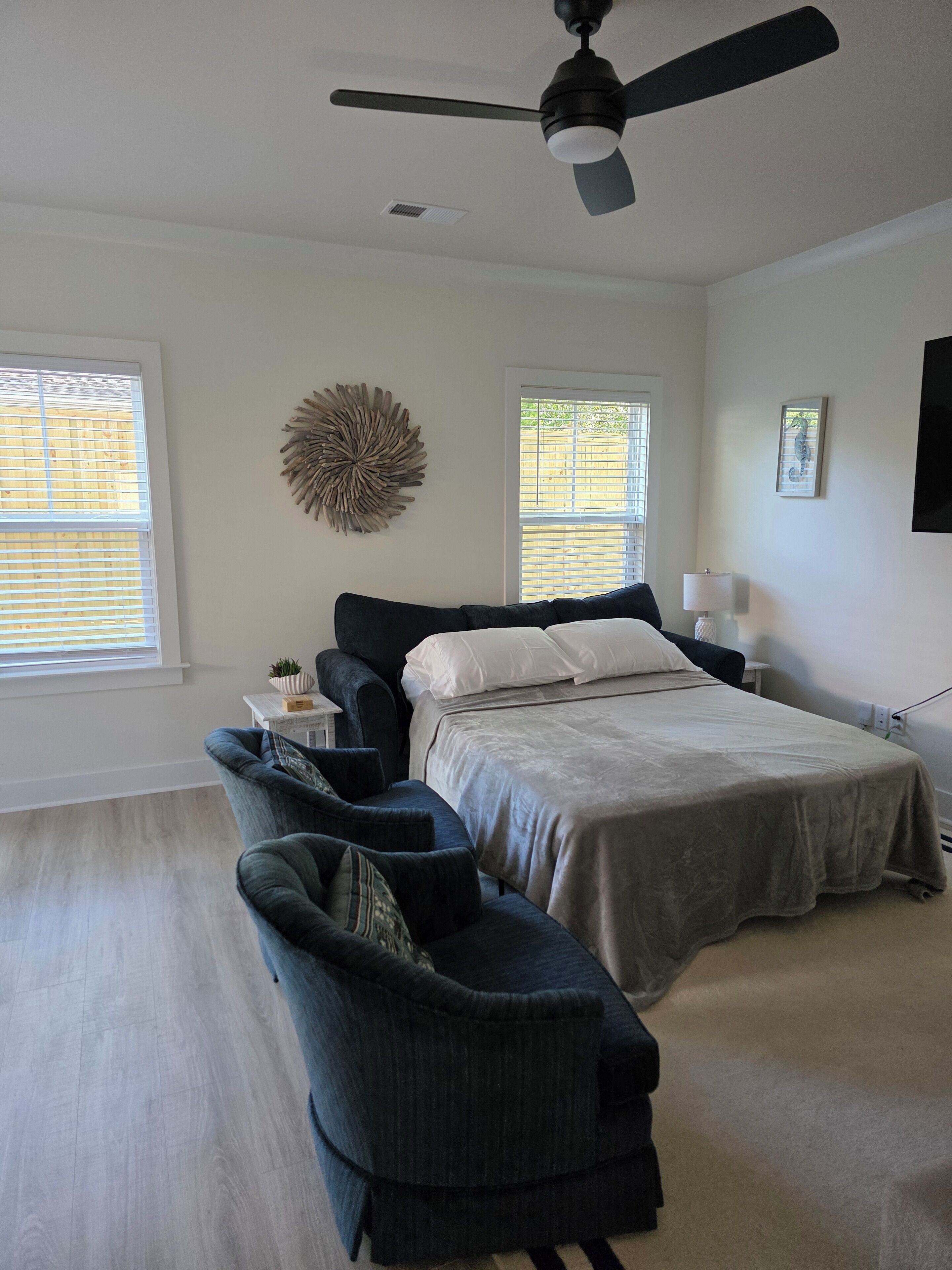 A bedroom with a bed, a ceiling fan, two windows, a wall art piece, a lamp, and a dark-colored sofa.