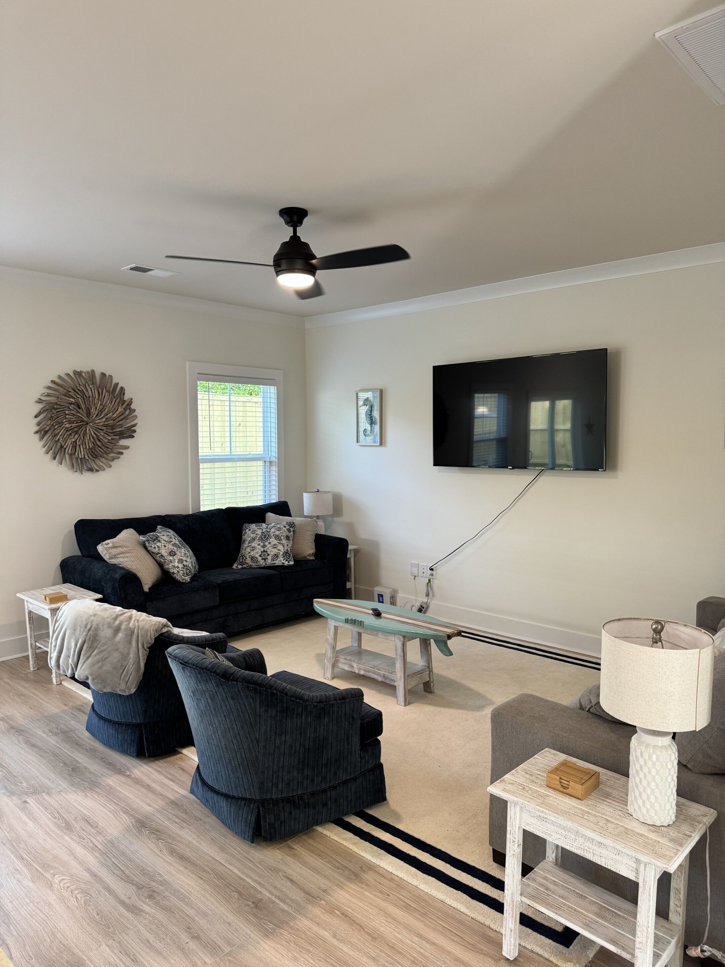 Ein Wohnzimmer mit Deckenventilator, einem an der Wand montierten Flachbildfernseher, einem dunkelgrauen Sofa, einem Couchtisch und einer Lampe auf einem Beistelltisch.