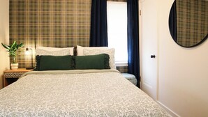 Un dormitorio con cama, mesita de noche, lámpara, planta en maceta, ventana con cortinas y espejo en la pared.