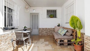 A patio with a stone floor, a white wall, a window with a plant, a sofa with cushions, and a dining area with chairs and a table. - Apartment 'Cristina' with Private Terrace, Wi-Fi, and Air Conditioning (Deltebre)
