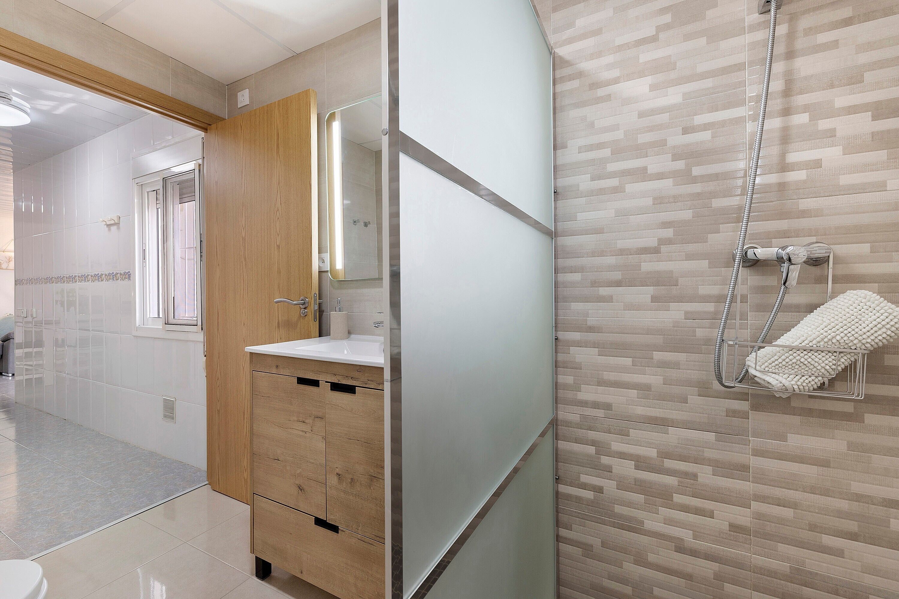 A modern bathroom with a glass shower enclosure, a wooden vanity, and a tiled floor.