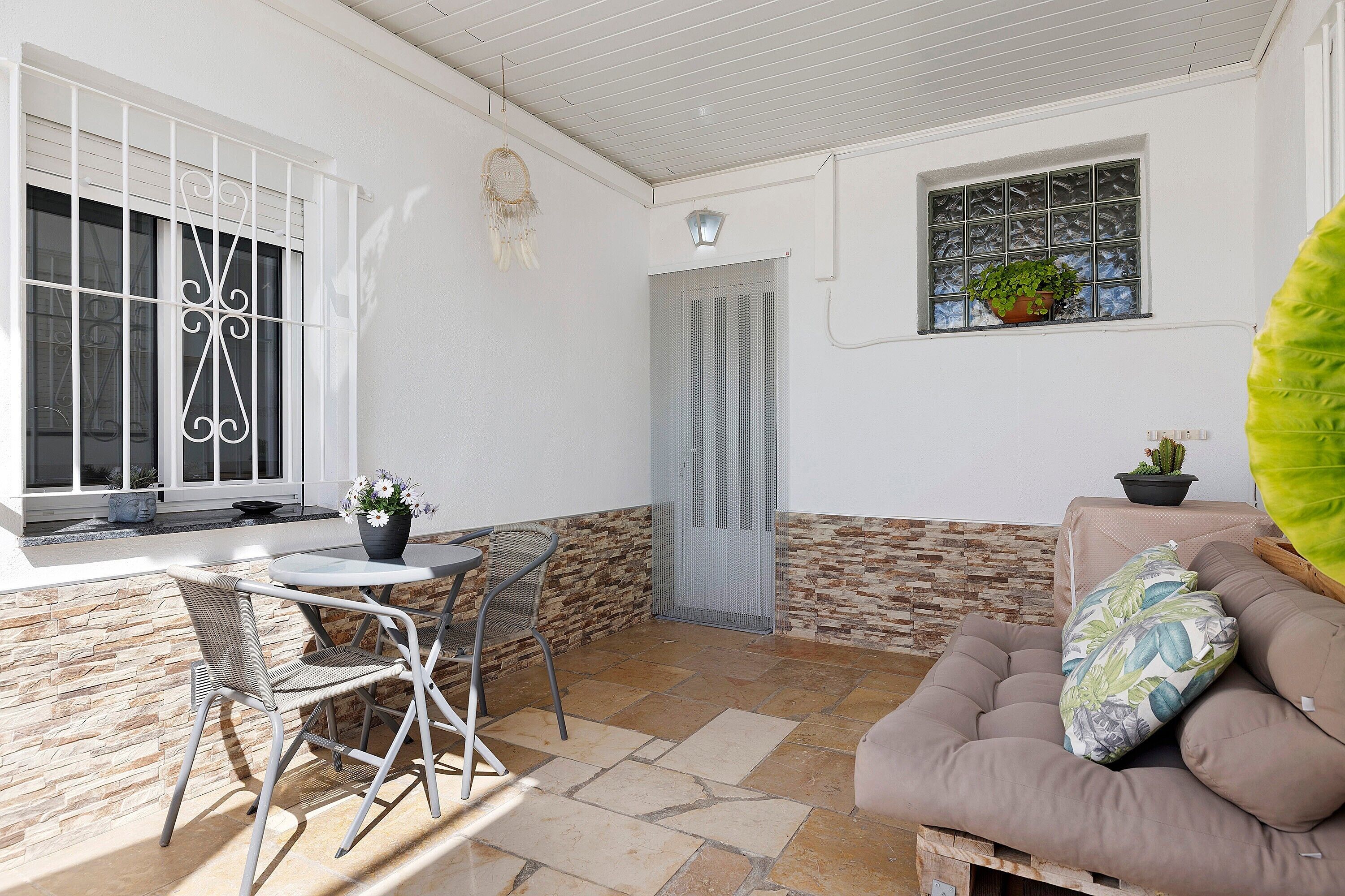 Un patio con una mesa y sillas, un área de descanso con cojines, una ventana con una planta y una puerta.