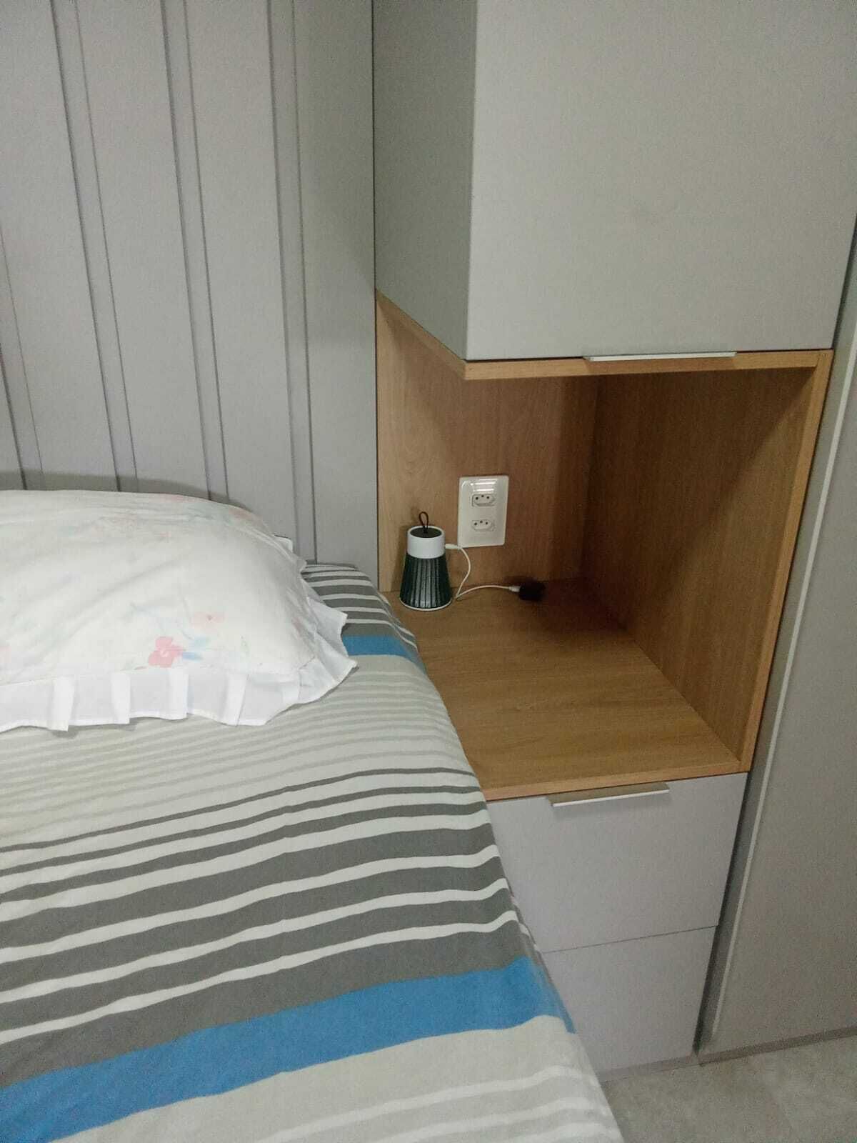 Sängyssä on raidallinen peitto, yöpöydällä on lamppu ja seinässä on pistorasia.