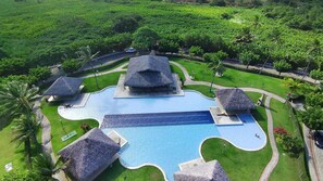 Ein großer Pool mit ungewöhnlicher Form, umgeben von strohgedeckten Gebäuden und üppiger Vegetation.