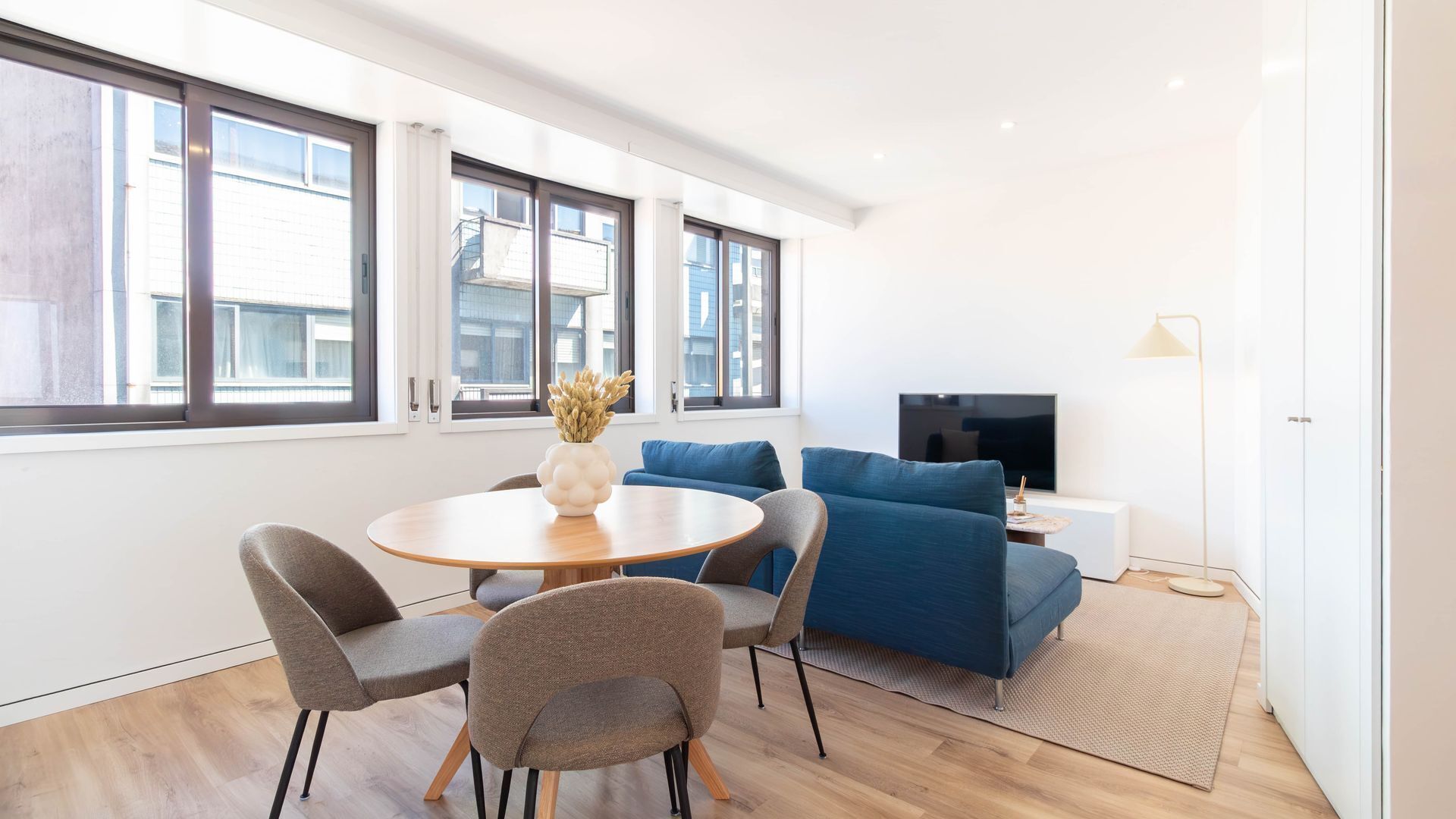 A modern living room with a round wooden table, grey and blue sofas, a flat-screen TV, and large windows.