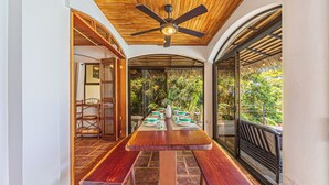 Uma área de refeições com ventoinha de madeira no teto, uma mesa grande com bancos e vista para o verde através de uma porta de vidro.