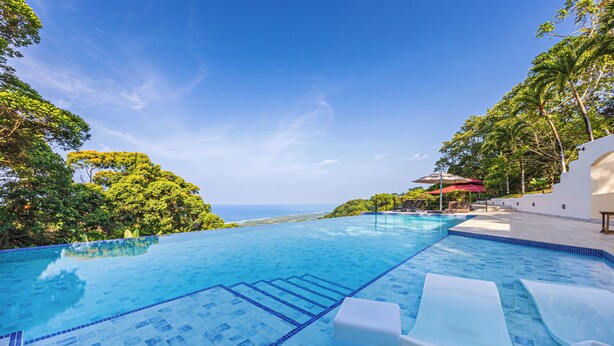 Uma piscina infinita azul cristalina com vista para o oceano, rodeada por vegetação exuberante e um edifício branco com um guarda-sol vermelho.