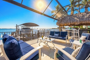 A sunlit outdoor seating area with a view of the sea, a covered boat, and a thatched roof structure. - VRCC Villa Pura Vida (Cape Coral)