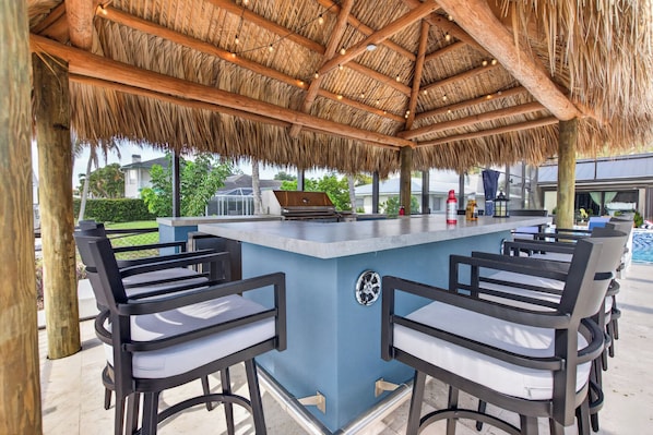 An outdoor bar area with a thatched roof, bar stools, and a grill. - VRCC Villa Pura Vida (Cape Coral)