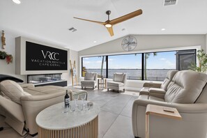 Ein geräumiges Wohnzimmer mit großem Fenster und Blick auf das Wasser, Deckenventilator und einem wandmontierten Flachbildfernseher.