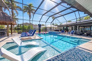 A swimming pool with lounge chairs and a hot tub, surrounded by a covered area with a view of palm trees and houses. - VRCC Villa Pura Vida (Cape Coral)