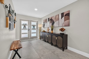 A hallway with a wooden bench, a glass door, a console table with decorative items, and abstract wall art. - VRCC Villa Pura Vida (Cape Coral)
