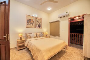 A bedroom with a bed, bedside tables, wall art, an air conditioning unit, and a balcony with a railing.
