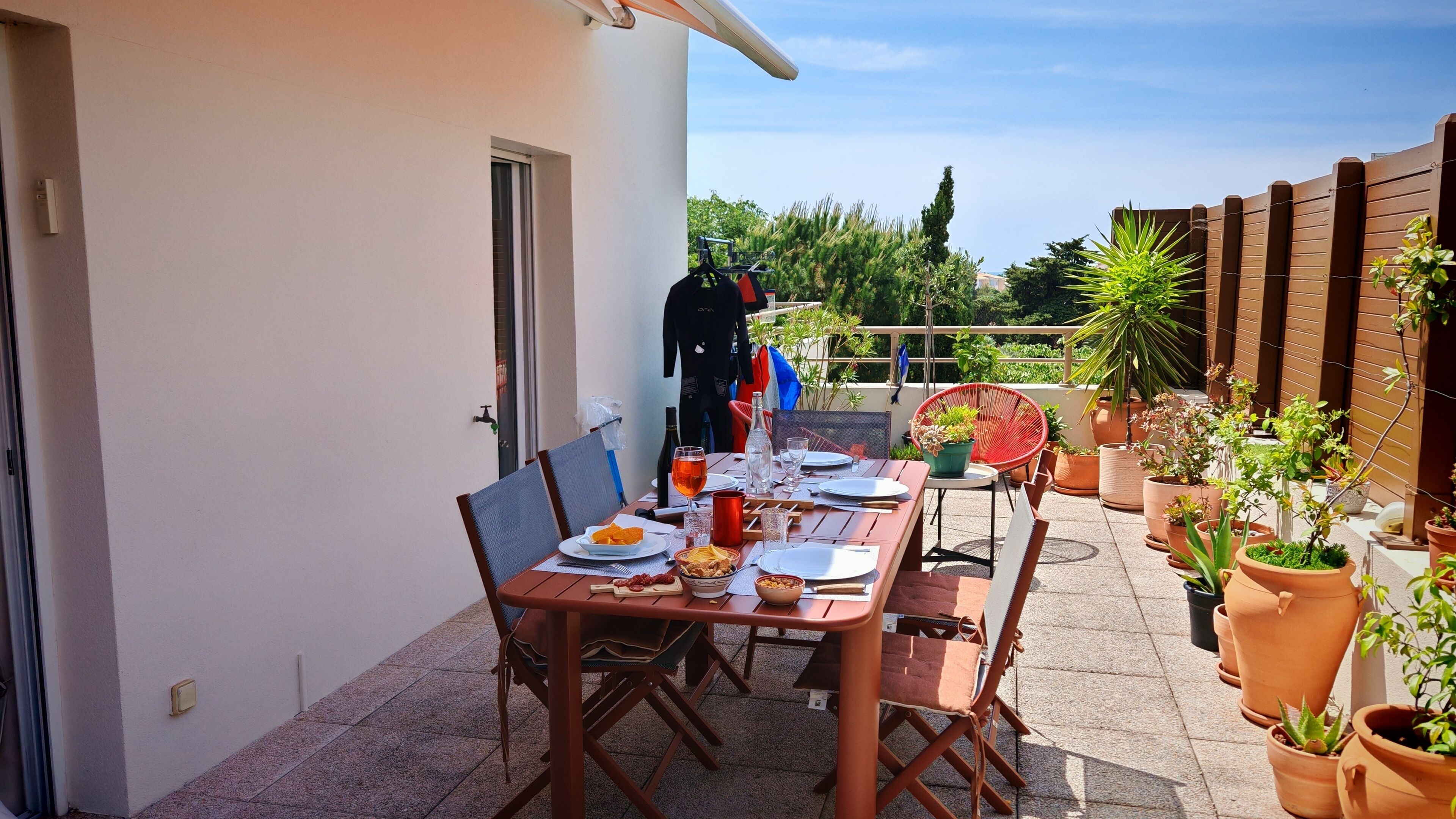 Un área de comedor al aire libre con una mesa lista para dos personas, rodeada de macetas con plantas y una valla de madera.