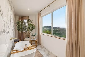 A bathroom with a bathtub, a potted plant, and a view of a landscape through a large window. - Hillside 2-Story | Dual Living | Mountain View (Vista)