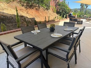 Ein Essbereich im Freien mit einem rechteckigen Tisch und Stühlen, umgeben von einem angelegten Garten mit Kakteen und Blick auf einen Hügel.