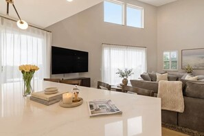 A modern living room with a flat-screen TV, a sofa, a dining table with a vase of flowers, and a magazine. - Hillside 2-Story | Dual Living | Mountain View (Vista)