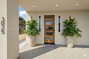 A building entrance with a wooden door, two potted plants, and a number '3025' on the wall.