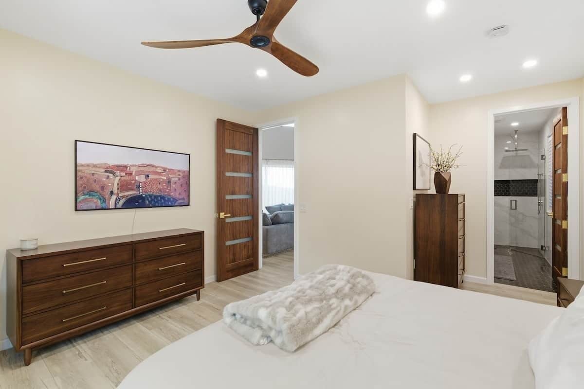 A bedroom with a bed, a wooden dresser, a ceiling fan, a painting, and a bathroom visible through an open door.