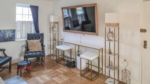 Una habitación con una butaca de cuero negro, un sofá blanco, una mesa de centro de vidrio, una lámpara de pie, un televisor montado en la pared y un estante decorativo con un reloj y figuritas.