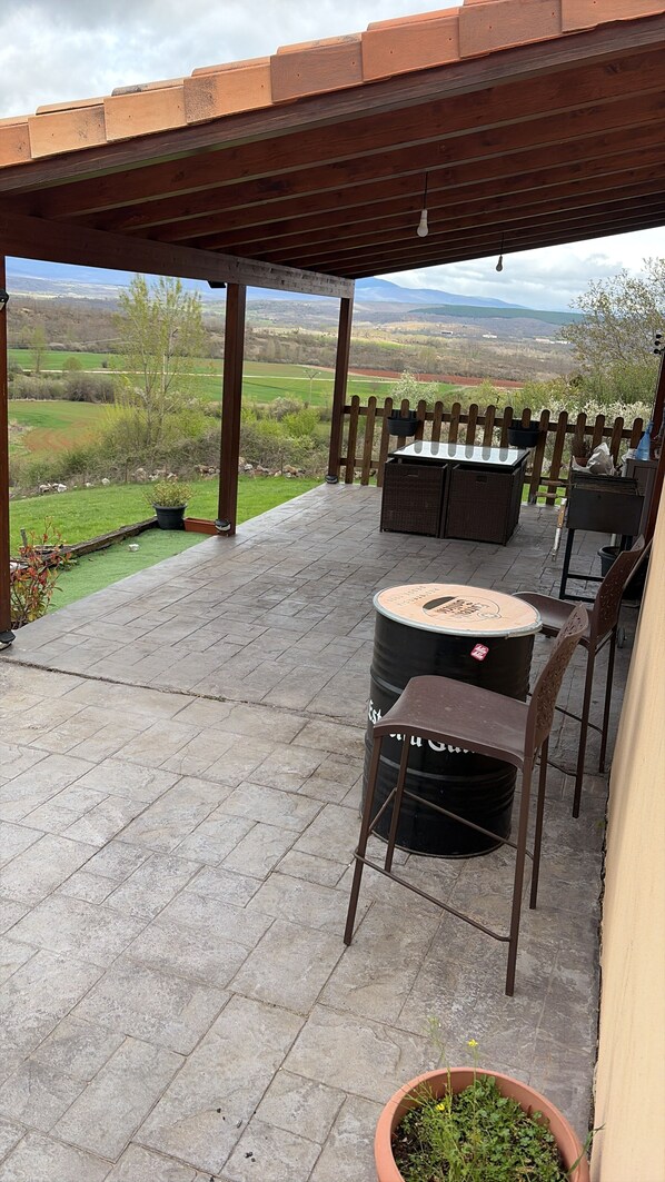 A covered patio with a view of the countryside, a table with chairs, and a barrel.