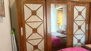 A bedroom with a wooden wardrobe, a bed with a pink bedspread, and a view into another room.