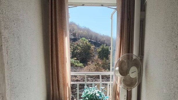 A room with a window, curtains, a fan, and a view of a hilly landscape.