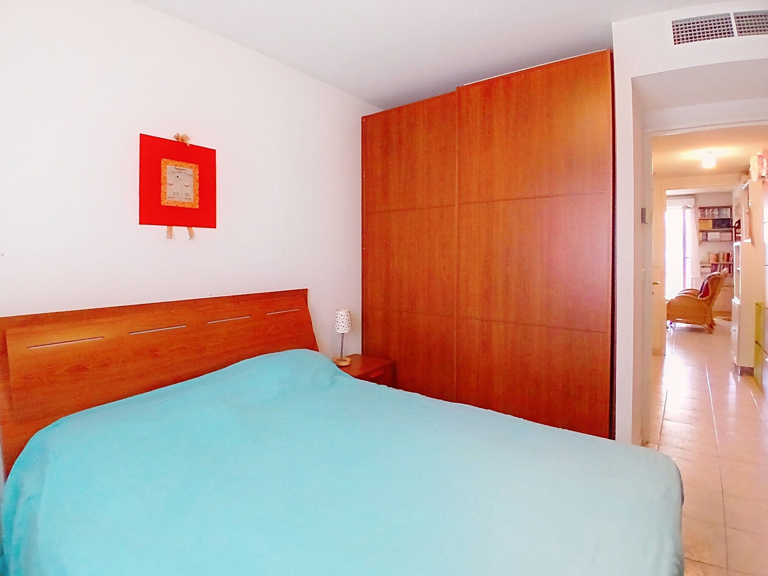 Une chambre à coucher avec une tête de lit en bois, un lit simple, une table de chevet, un tableau encadré de rouge et une armoire.