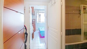 Een smalle gang met een houten garderobe, een deur naar een badkamer met douche, en een kijkje in een slaapkamer met bed en raam.