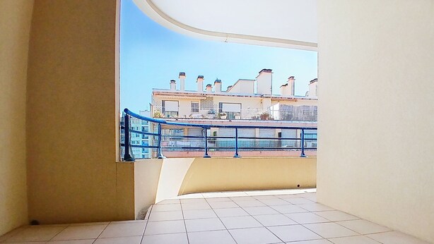 Een balkon met betegelde vloer, een blauwe balustrade en een uitzicht op gebouwen in de verte.