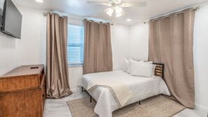 Quarto com cama, cômoda de madeira, ventilador de teto e televisão fixada na parede.