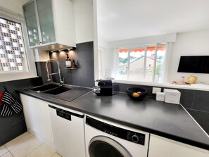 A modern kitchen with a dishwasher, double sink, and a window with a view of buildings. - Central Antibes, Spacious and Bright 2-Bedroom Apartment Near Beaches (Antibes)