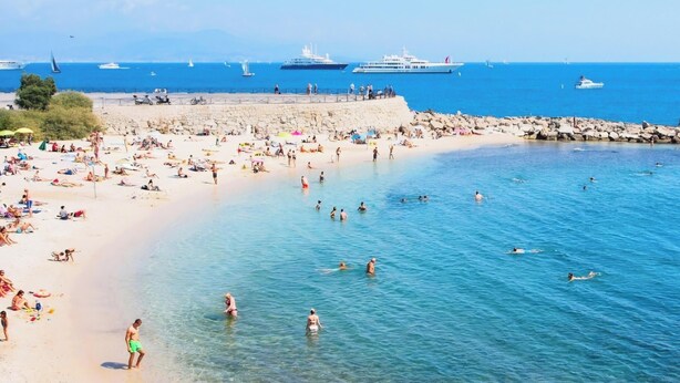 En strand med folk som badar och solar, båtar på vattnet och en stenbrytning som fungerar som vågbrytare.