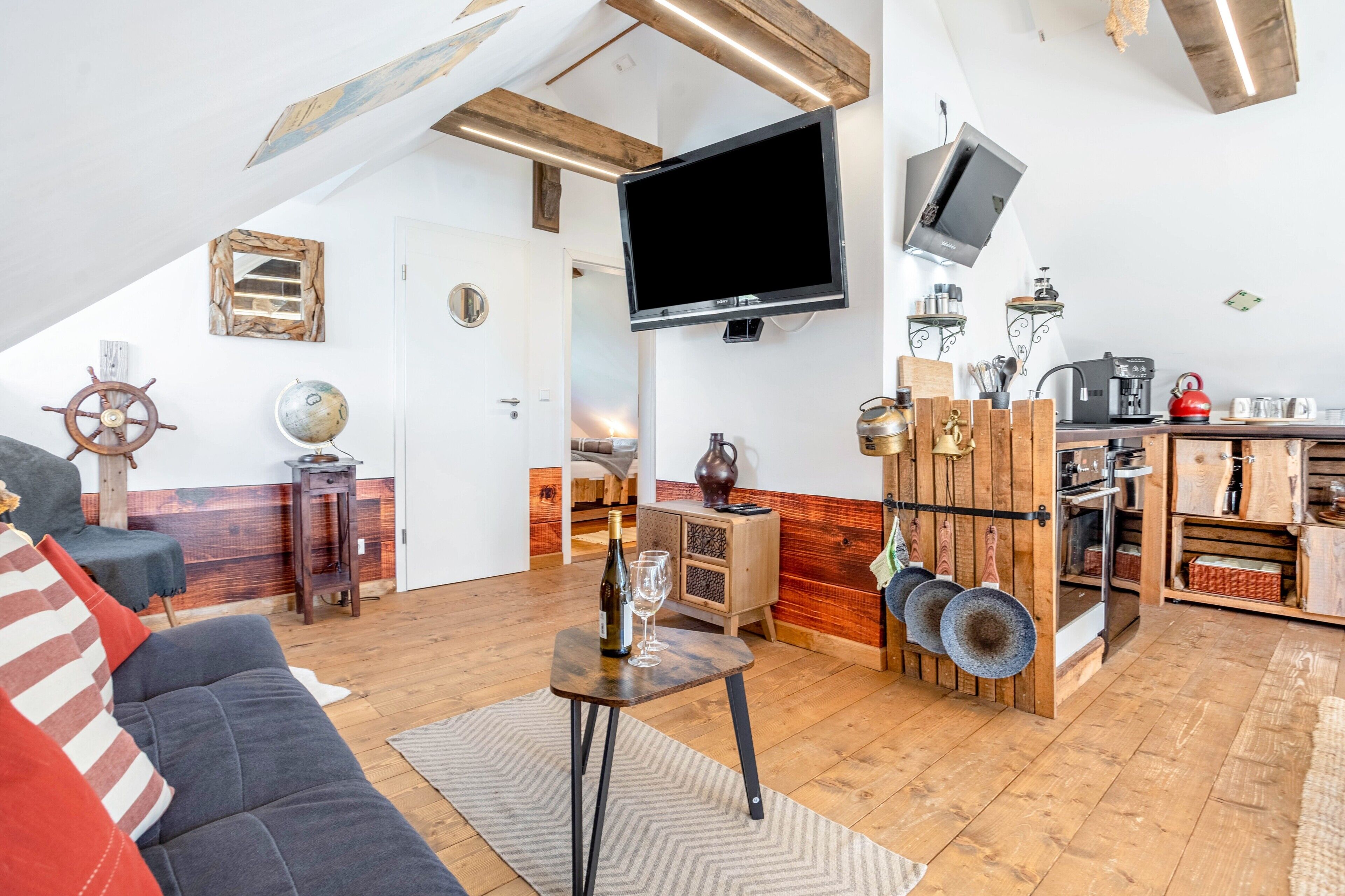 Un salon confortable avec un canapé, une petite table sur laquelle se trouvent une bouteille et un verre, un téléviseur fixé au mur, et un coin cuisine doté de meubles en bois et d’une machine à café.