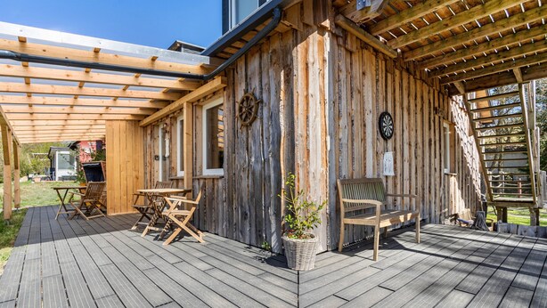 Eine Holzterrasse mit Gartenmöbeln, einer Pergola und einem rustikalen Gebäude mit Schiffsschwengel.