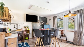 Eine kleine Küche mit modernen Geräten, ein Essbereich mit Stühlen, ein an der Wand montierter Fernseher und ein Fenster mit Vorhängen.