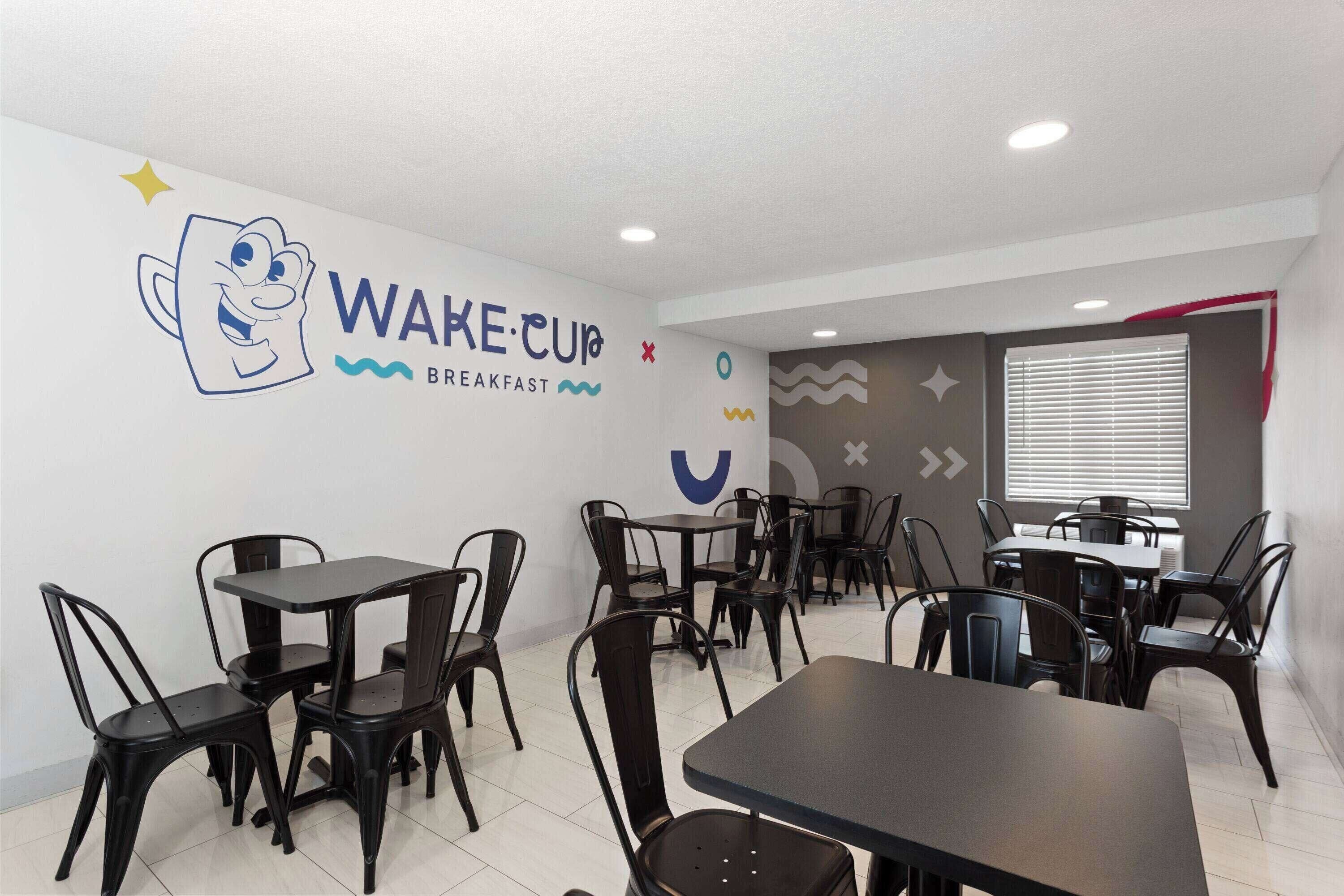 Cafetería para desayunos con sillas y mesas de metal negro, una pared con un personaje de caricatura y el texto 'WAKE CUP BREAKFAST'.
