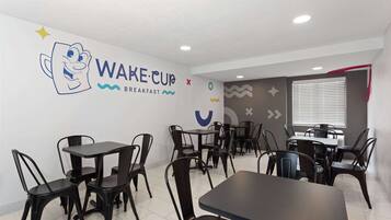 Cafetería para desayunos con sillas y mesas de metal negro, una pared con un personaje de caricatura y el texto 'WAKE CUP BREAKFAST'.