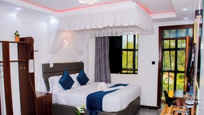 A bedroom with a bed, a nightstand, a wardrobe, a window with curtains, and a view of greenery outside. - The Grand Brystal Hotel (Busia)