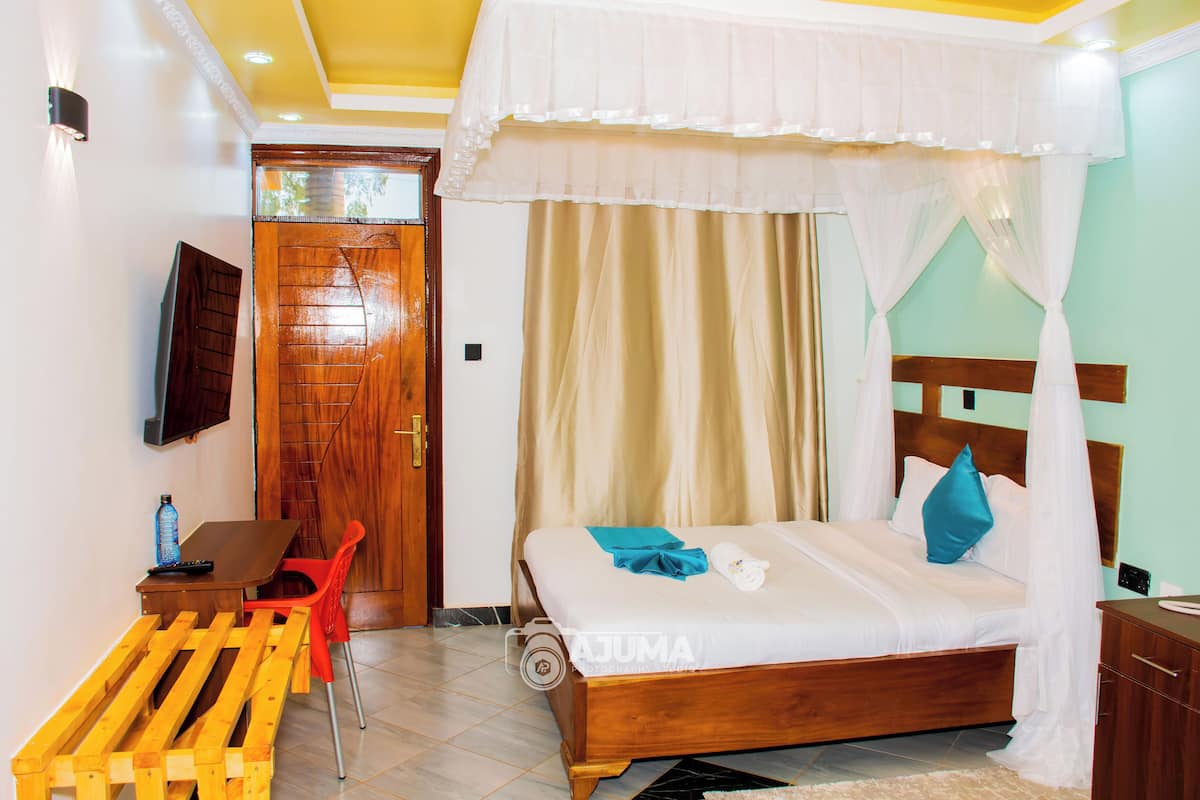 A bedroom with a wooden bed, a desk with a chair, a TV mounted on the wall, and a door with a wooden panel.