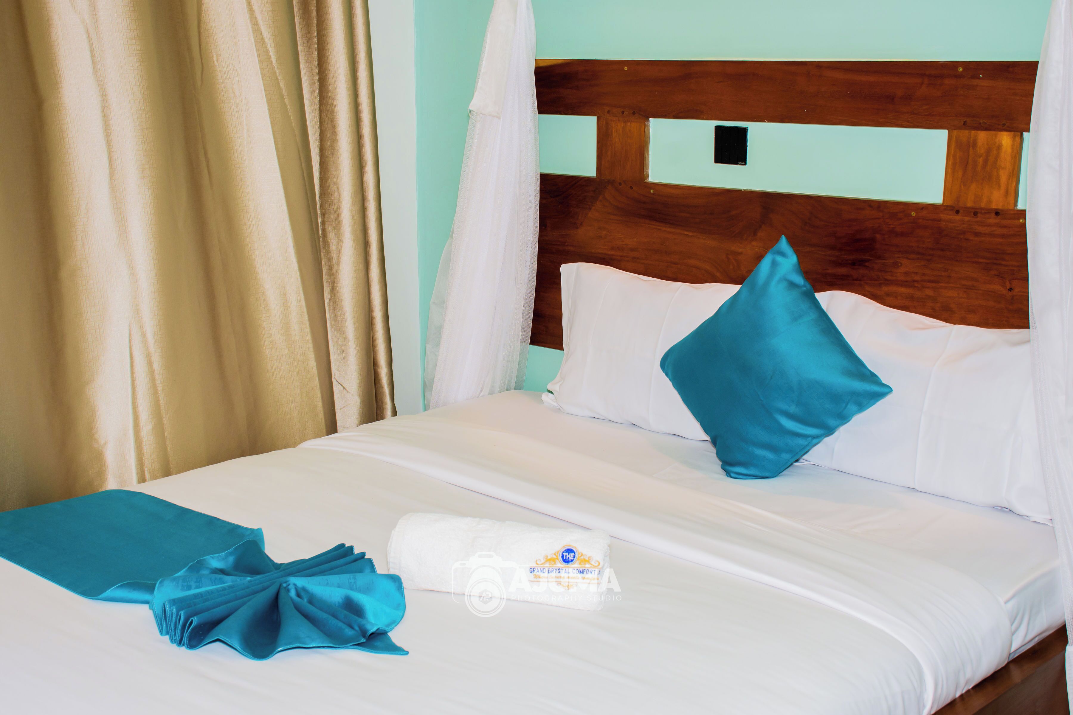 A bed with white bedding, a teal throw blanket, and a white towel with a logo.