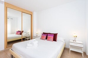 Ein Hotelzimmer mit großem Spiegel, einem Bett mit weißer Bettwäsche, farbenfrohen Kissen, einem Nachttisch mit Lampe und einem Beistelltisch ebenfalls mit Lampe.