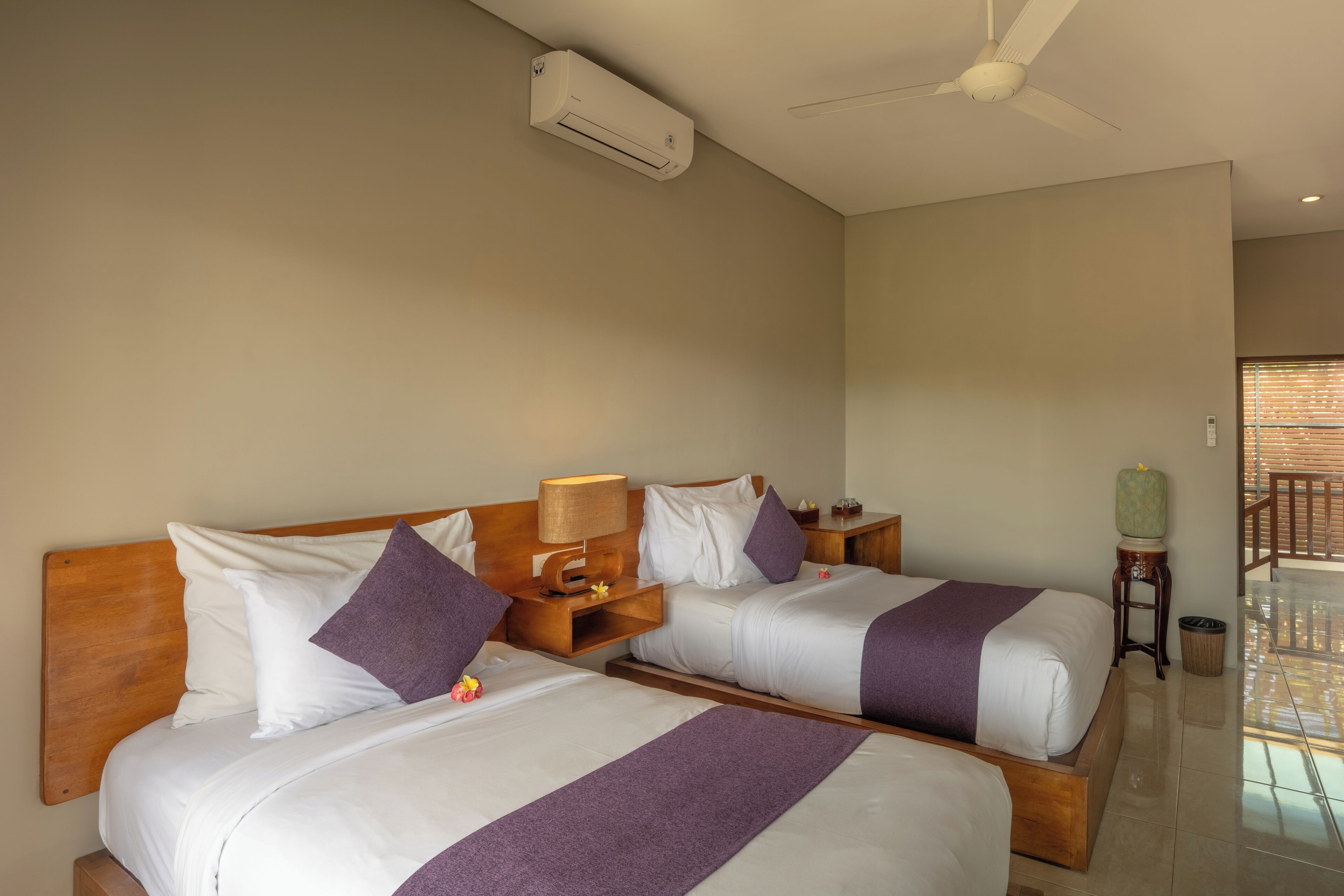 Habitación de hotel con dos camas, ventilador de techo, unidad de aire acondicionado y vistas al exterior.